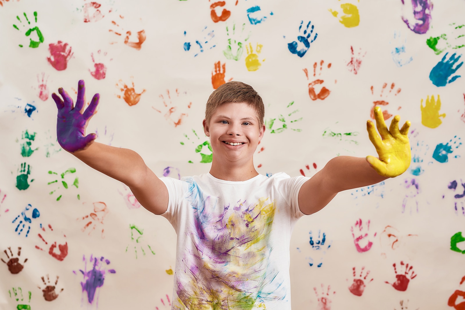 Happy disabled boy with Down syndrome smiling at camera while reaching out his hands painted in colorful paints ready for hand prints on the wall. Children with disabilities and special needs concept